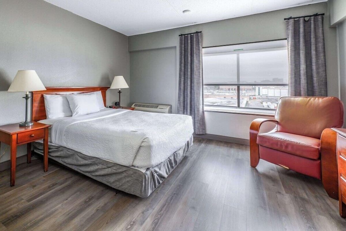 A hotel room with a bed, two bedside tables with lamps, a chair, a window with curtains, and a view of the city.