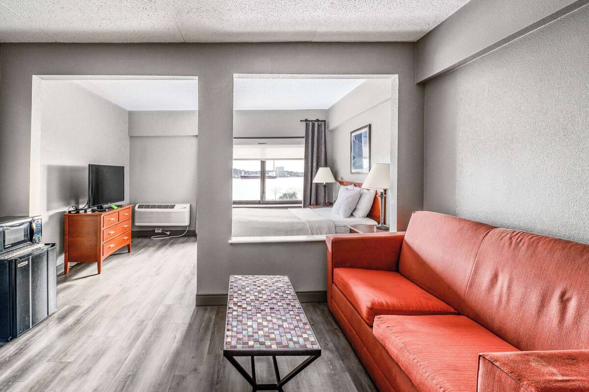 A modern hotel room with a red sofa, a small table, a TV, a dresser, and a window with a city view.