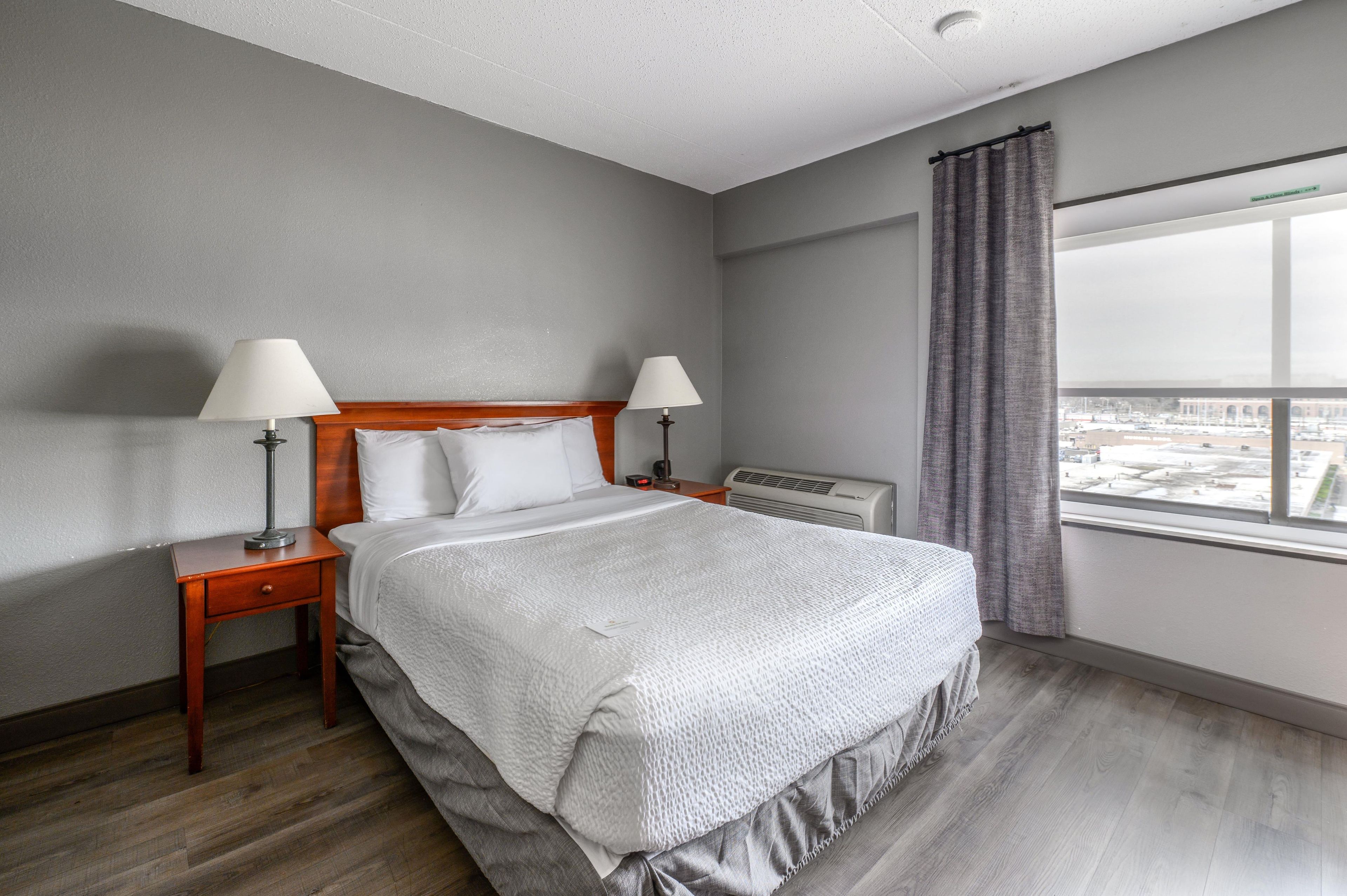 Ein Hotelzimmer mit einem Bett, zwei Nachttischlampen, einem Nachttisch, einem Fenster mit Vorhängen und Blick auf die Stadt.