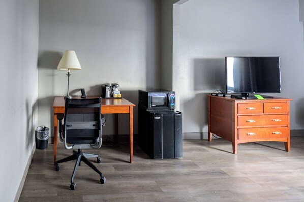 A hotel room with a wooden desk, a black office chair, a lamp, a microwave, a television on a wooden dresser, and a trash can.