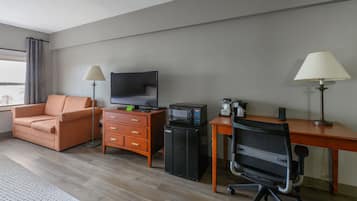 A hotel room with a sofa, a desk with a computer, a television, a coffee machine, and a chair.