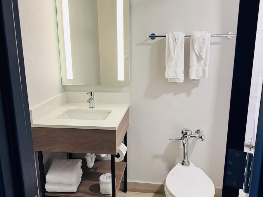 A modern bathroom with a sink, mirror, towel rack, toilet, and a wooden shelf.