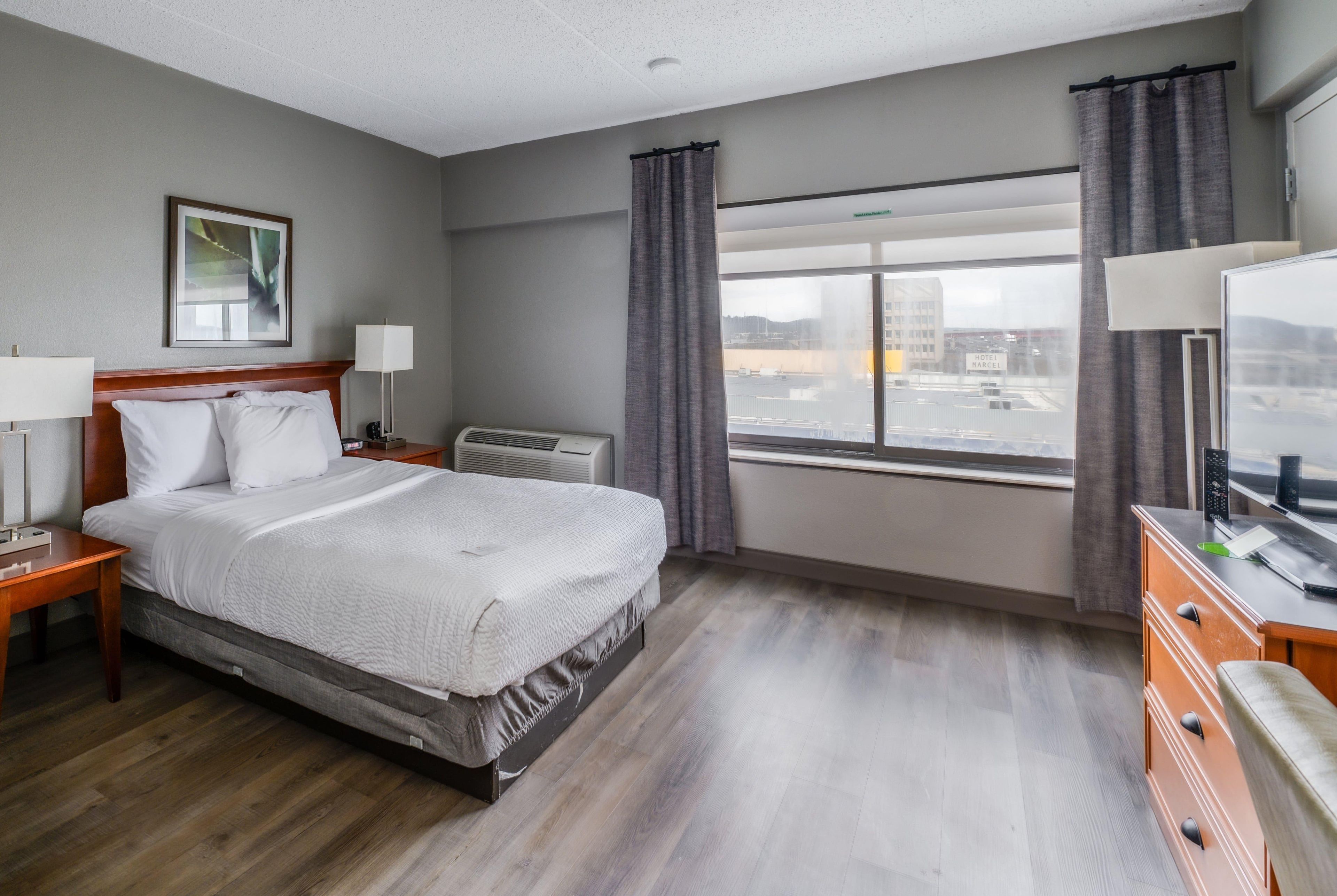 A hotel room with a bed, bedside tables, a desk, a chair, a window with curtains, and a view of the cityscape.