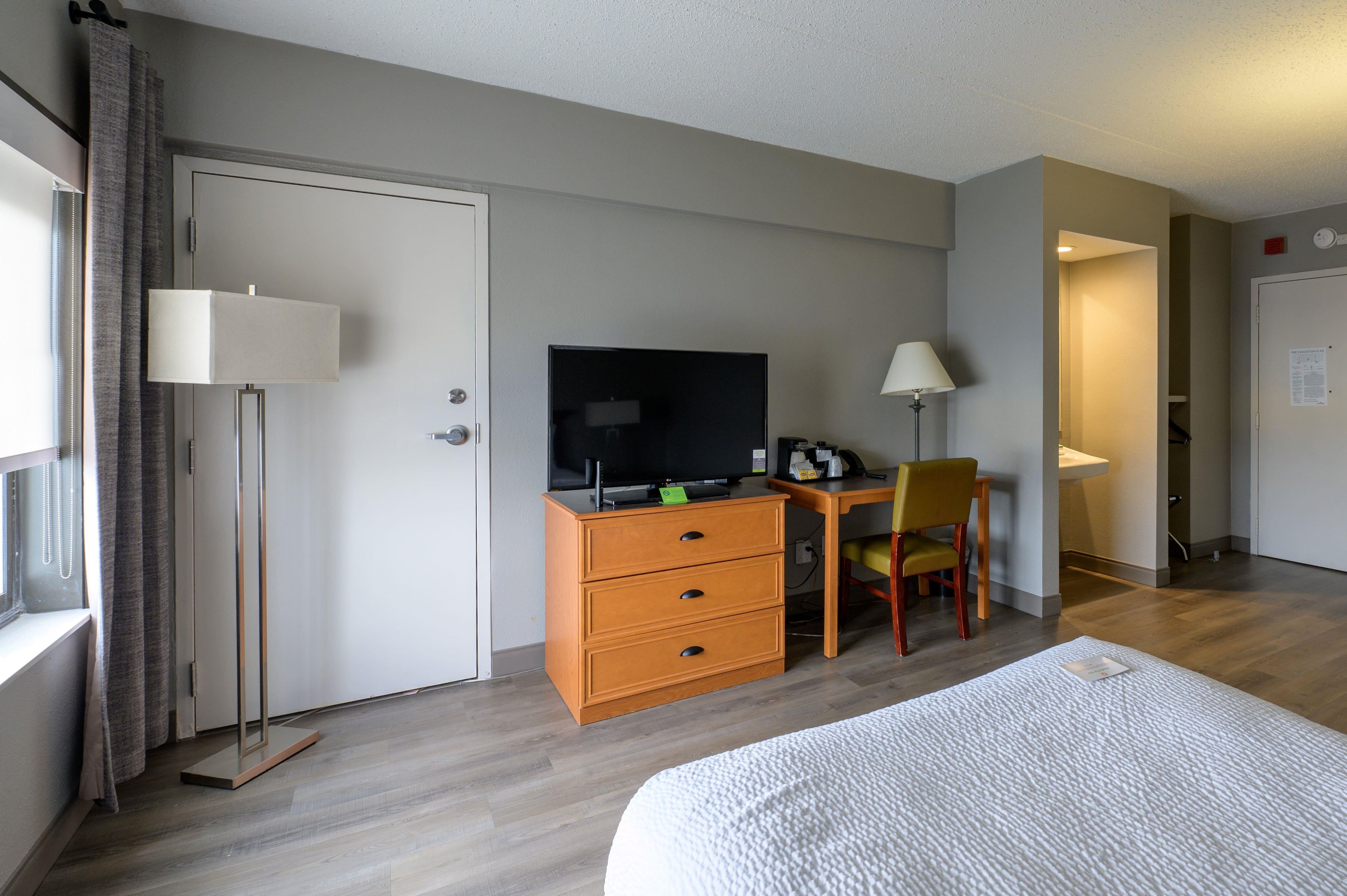 Una habitación de hotel con una cama, un escritorio con computadora, una silla, una lámpara y una puerta.