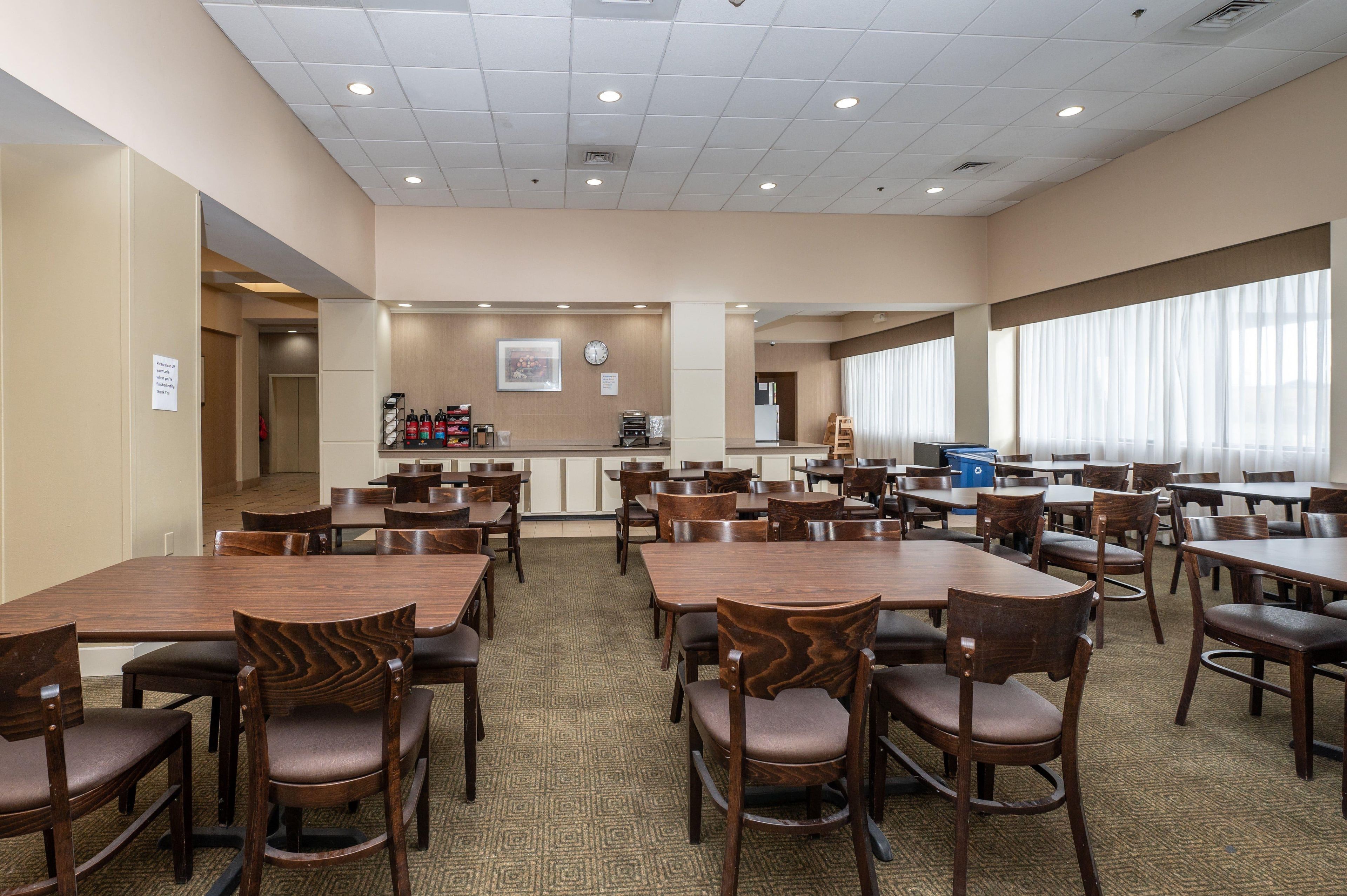 A hotel dining area with multiple tables and chairs, a clock, and a view of the outside through large windows.