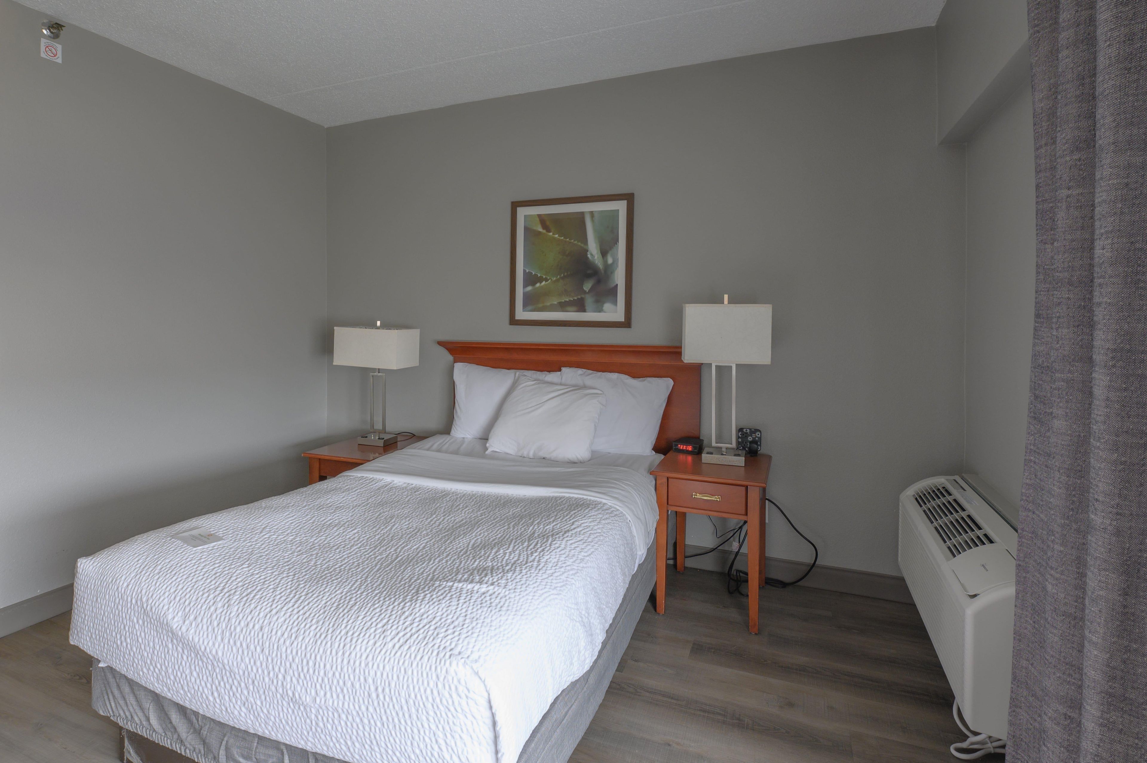 Habitación de hotel con cama, dos mesitas de noche, una mesita auxiliar, un reloj de pared, un cuadro enmarco y un calentador portátil.