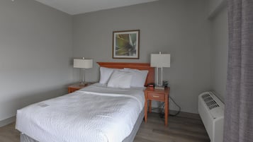 A hotel room with a bed, two bedside tables, a nightstand, a wall-mounted clock, a framed picture, and a standing heater.