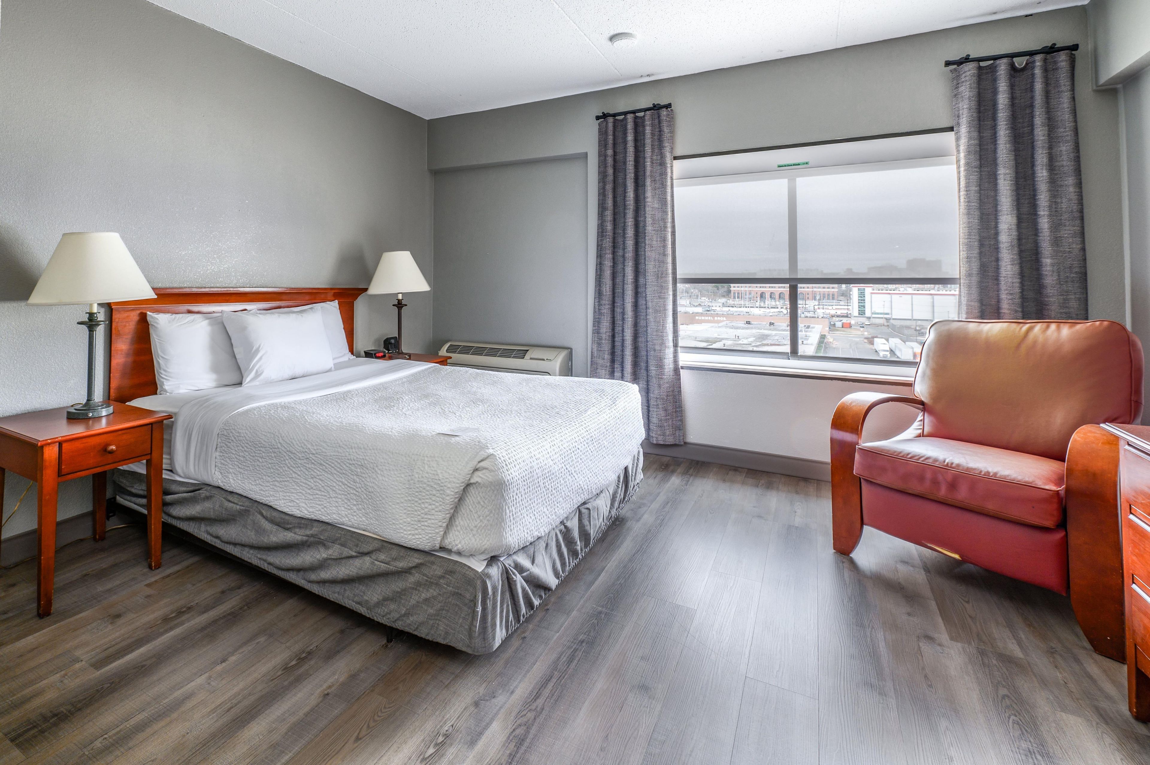 A hotel room with a bed, two bedside tables with lamps, a chair, a window with curtains, and a view of the airport.