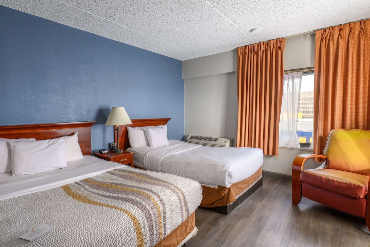 A hotel room with two beds, a nightstand with a lamp, an air conditioning unit, a window with curtains, and a leather armchair.
