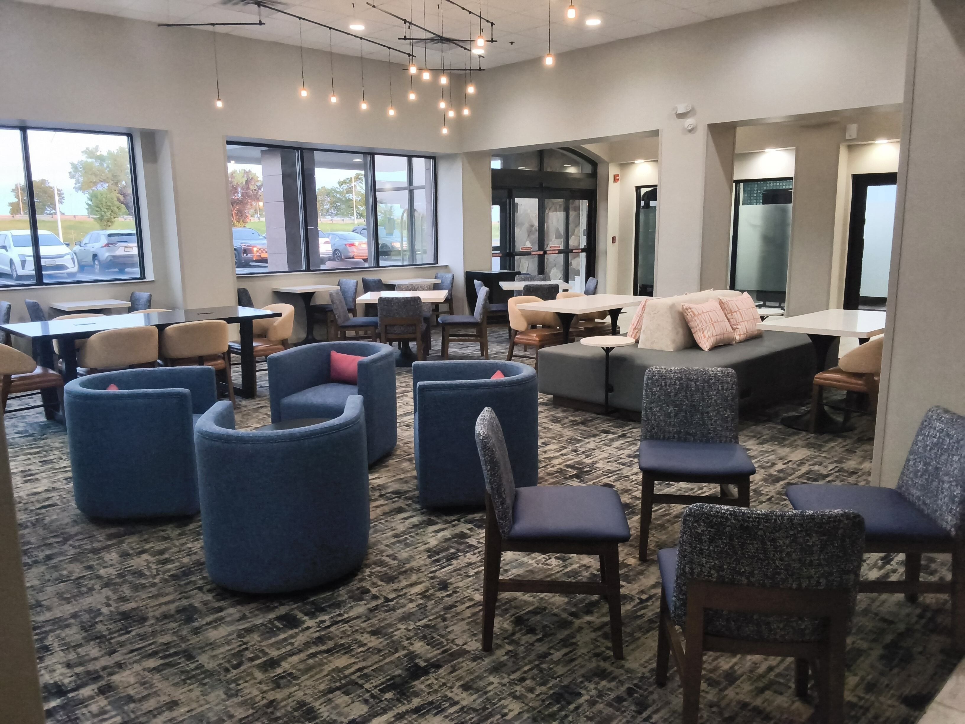 A modern lounge area with seating arrangements, large windows, and a carpeted floor.