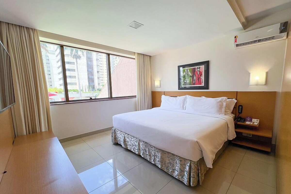 Habitación de hotel con una cama grande, un escritorio, una ventana con cortinas, un cuadro en la pared y un equipo de aire acondicionado.