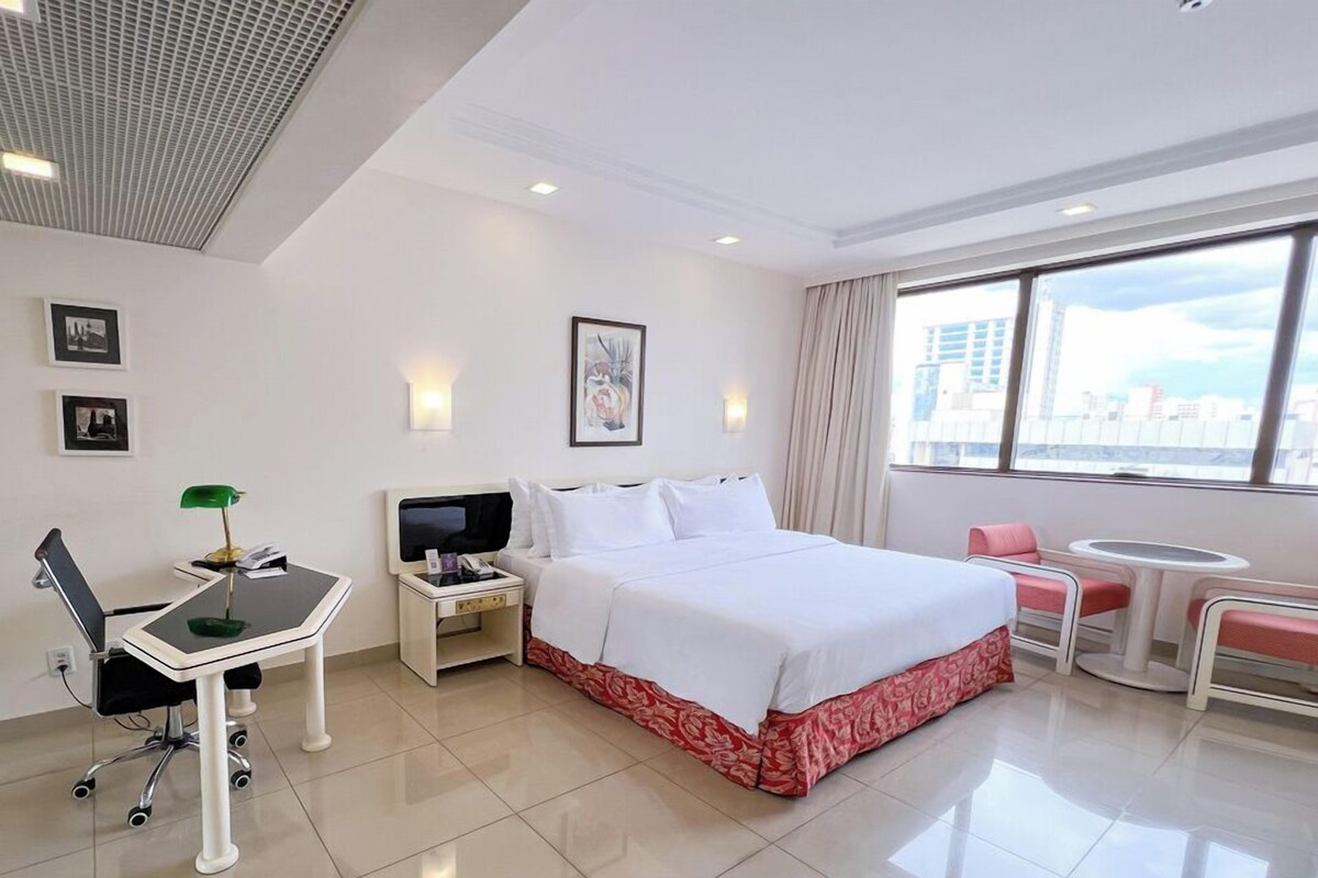 Habitación de hotel con cama, escritorio, silla, mesita de noche y ventana con vista a la ciudad.