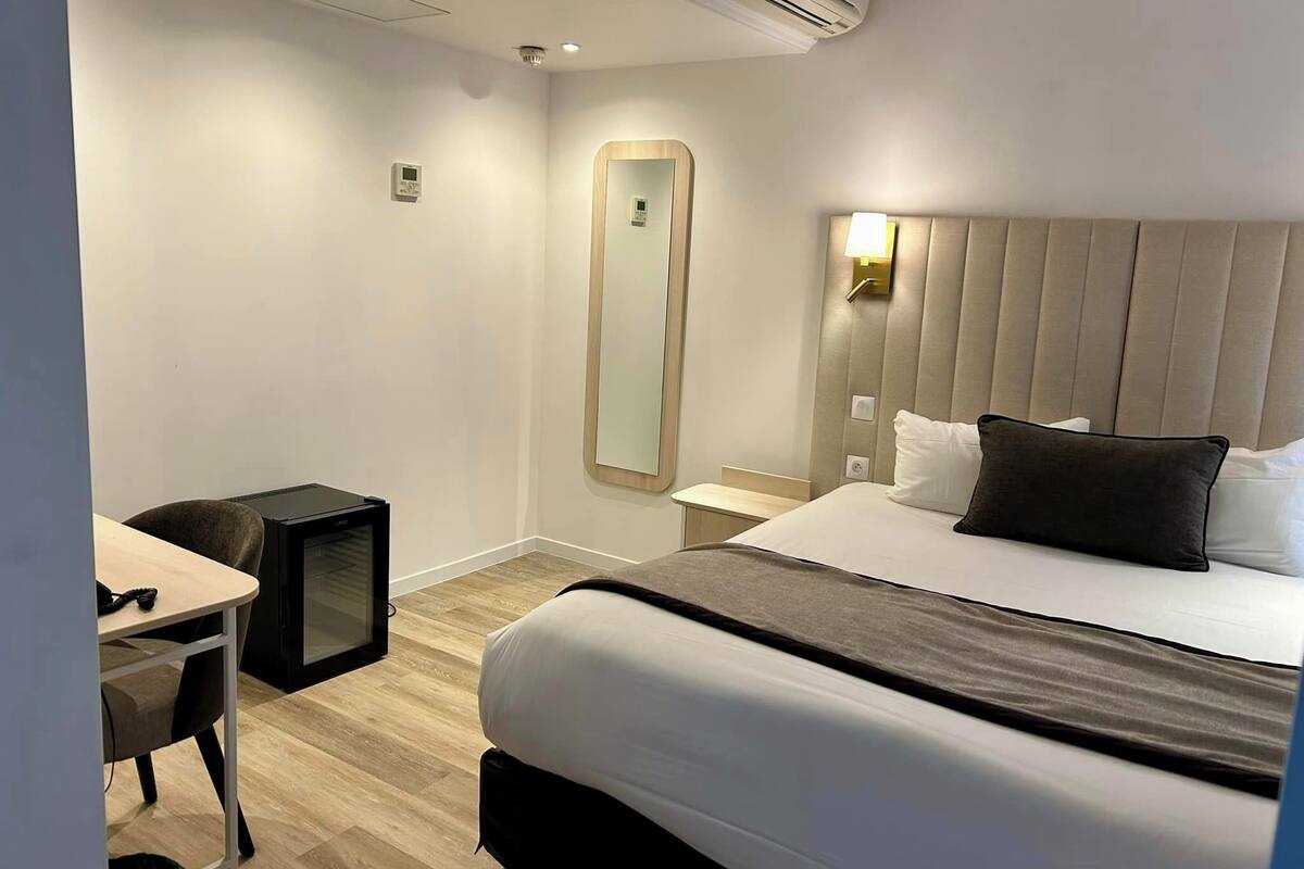 A hotel room with a bed, bedside table, lamp, mirror, desk, chair, and an air conditioning unit.