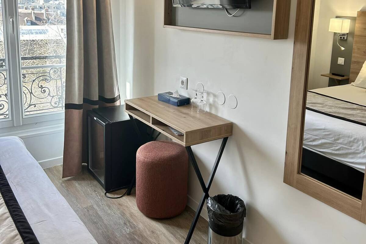 A hotel room with a bed, a desk with a computer, a TV mounted on the wall, a window with curtains, and a balcony view.
