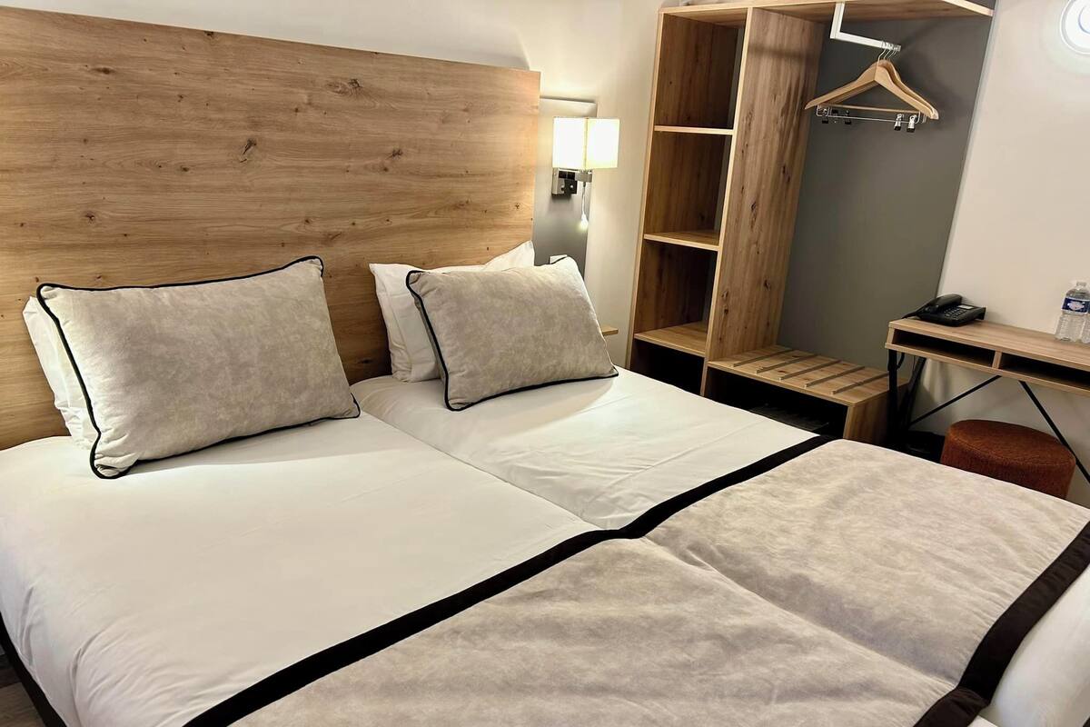A double bed with two pillows, a wooden headboard, a desk with a chair, and a hanging clothes rack.