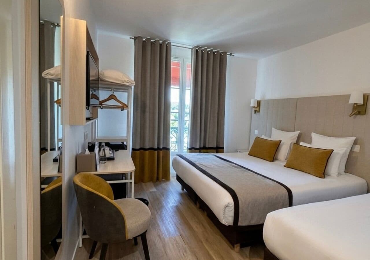 A hotel room with a bed, a desk with a chair, a wardrobe, and a window with curtains.