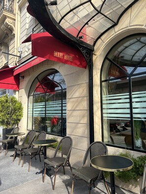 An outdoor seating area with wicker chairs and tables, a red awning with "Derby Hotel" signage, and large arched windows reflecting the sky. - Hotel Derby Eiffel (Paris)