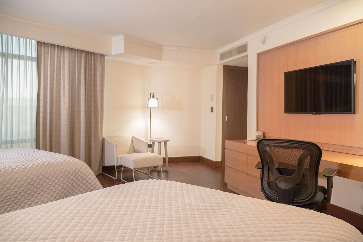 A hotel room with two beds, a desk, a chair, a lamp, and a television.