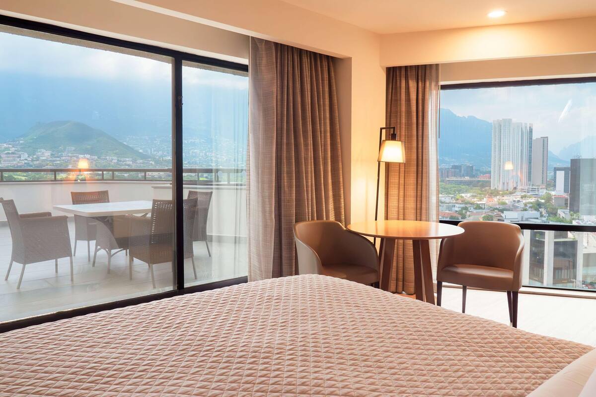 Quarto de hotel com uma cama grande, varanda com mesa e cadeiras e vista para a paisagem urbana.