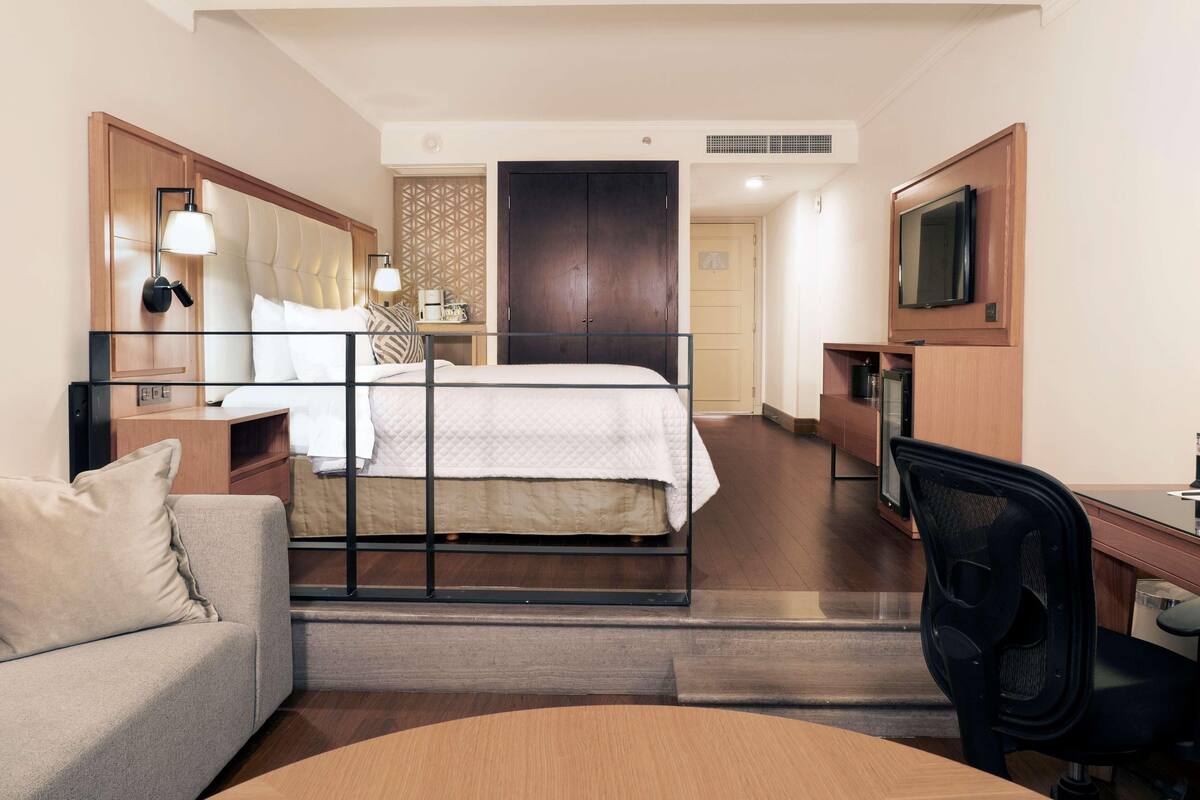 Quarto de hotel com uma cama, uma escrivaninha, uma cadeira, um sofá, uma televisão e um guarda-roupa.