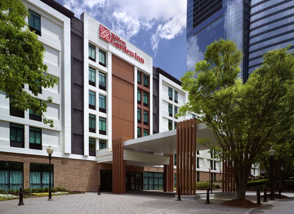 Un bâtiment moderne, reconnaissable à l’enseigne « Hilton Garden Inn », qui présente une combinaison de façades blanches et brunes, de grandes fenêtres et un espace d’entrée abrité agrémenté d’arbres.