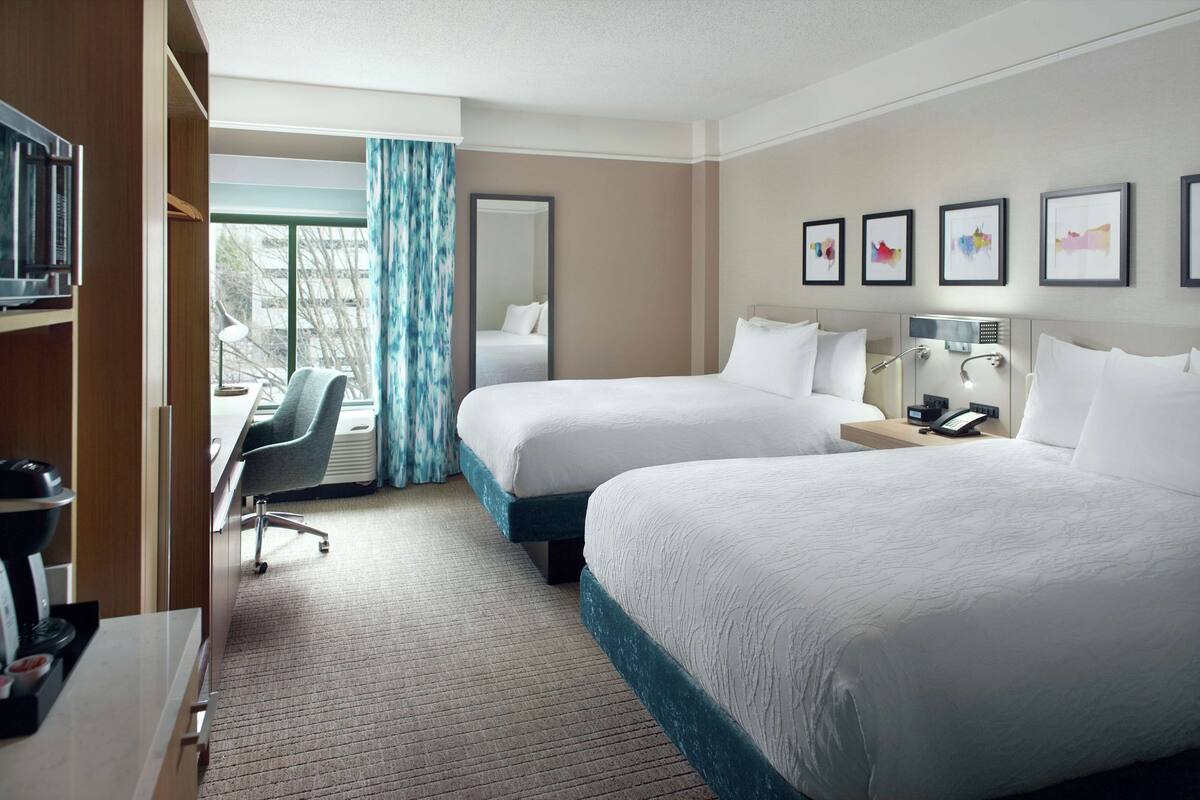 A hotel room with two beds, a desk, a chair, a desk lamp, a telephone, and framed pictures on the wall.