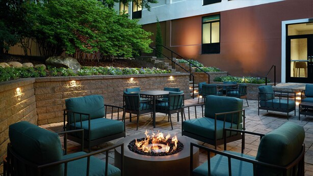 An outdoor patio with a circular fire pit surrounded by chairs and tables, a stone wall, and a staircase leading to an upper level.