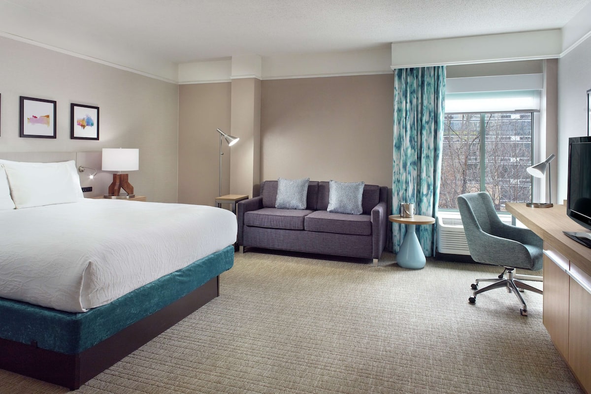 A hotel room with a large bed, a sofa, a desk with a chair, and a television.