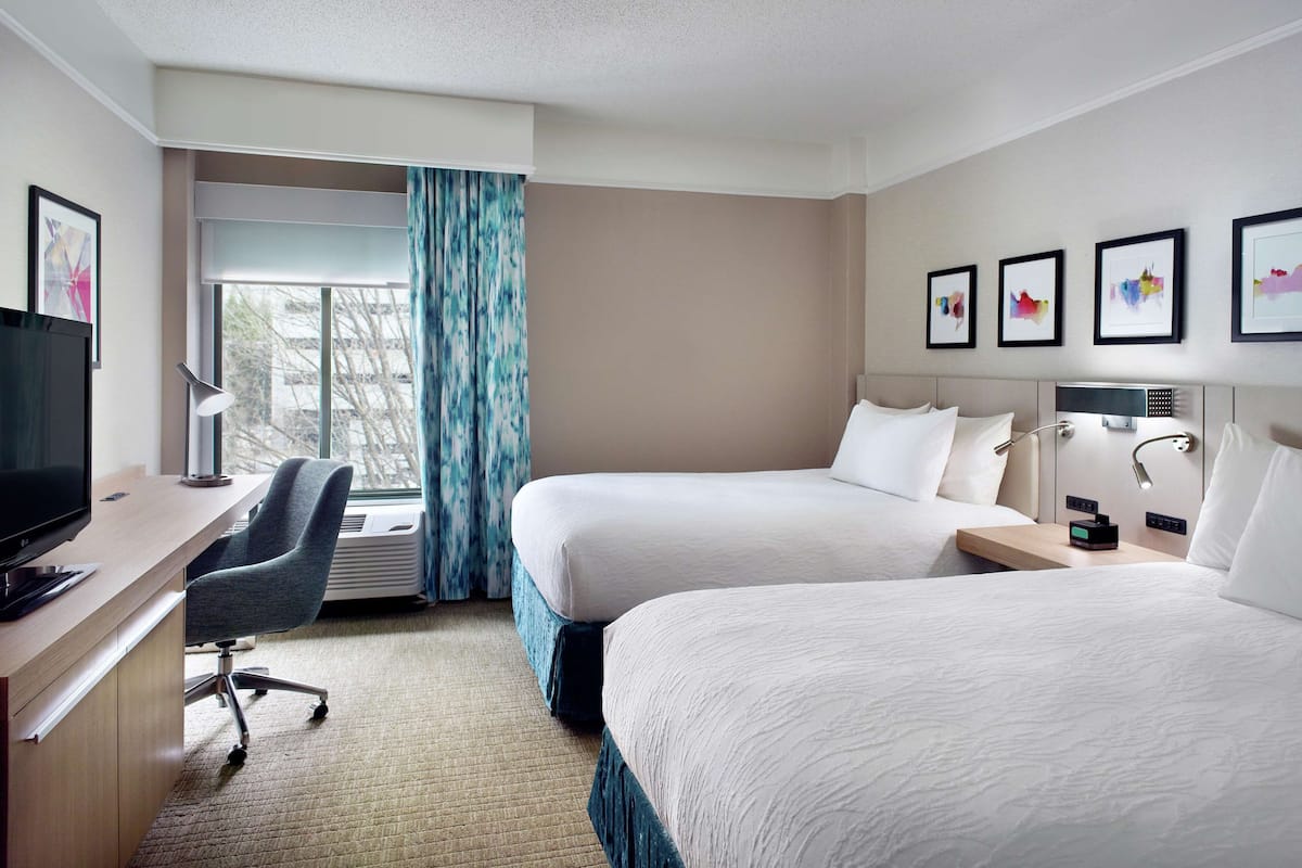 A hotel room with two beds, a desk with a chair, a TV, and a window with curtains.