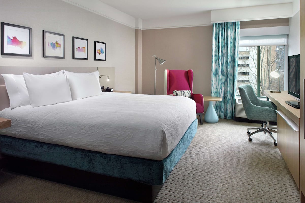 A hotel room with a large bed, a desk with a chair, a TV, and framed artwork on the wall.