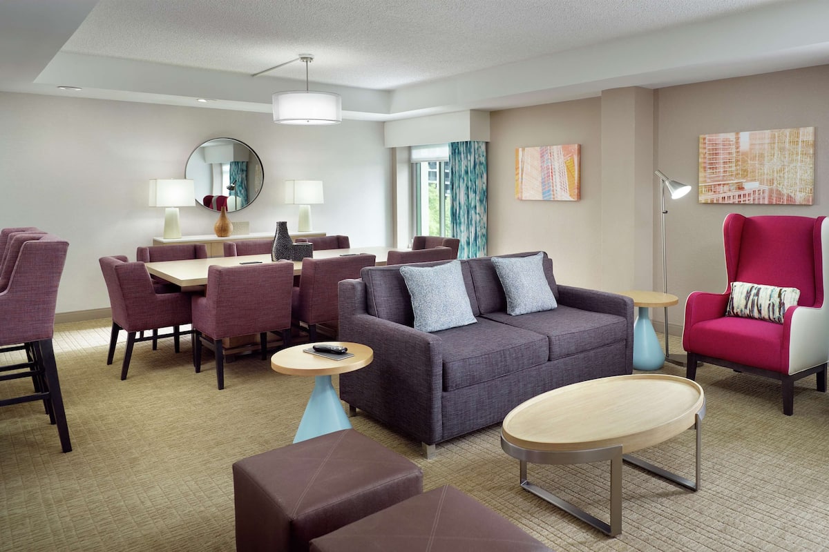 A modern hotel room with a dining area, a sofa set, and a large window.