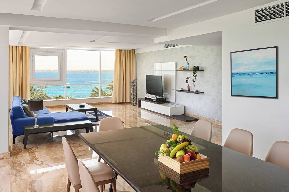 Habitación de hotel moderna con zona de comedor, sofá, televisor y vistas al mar.