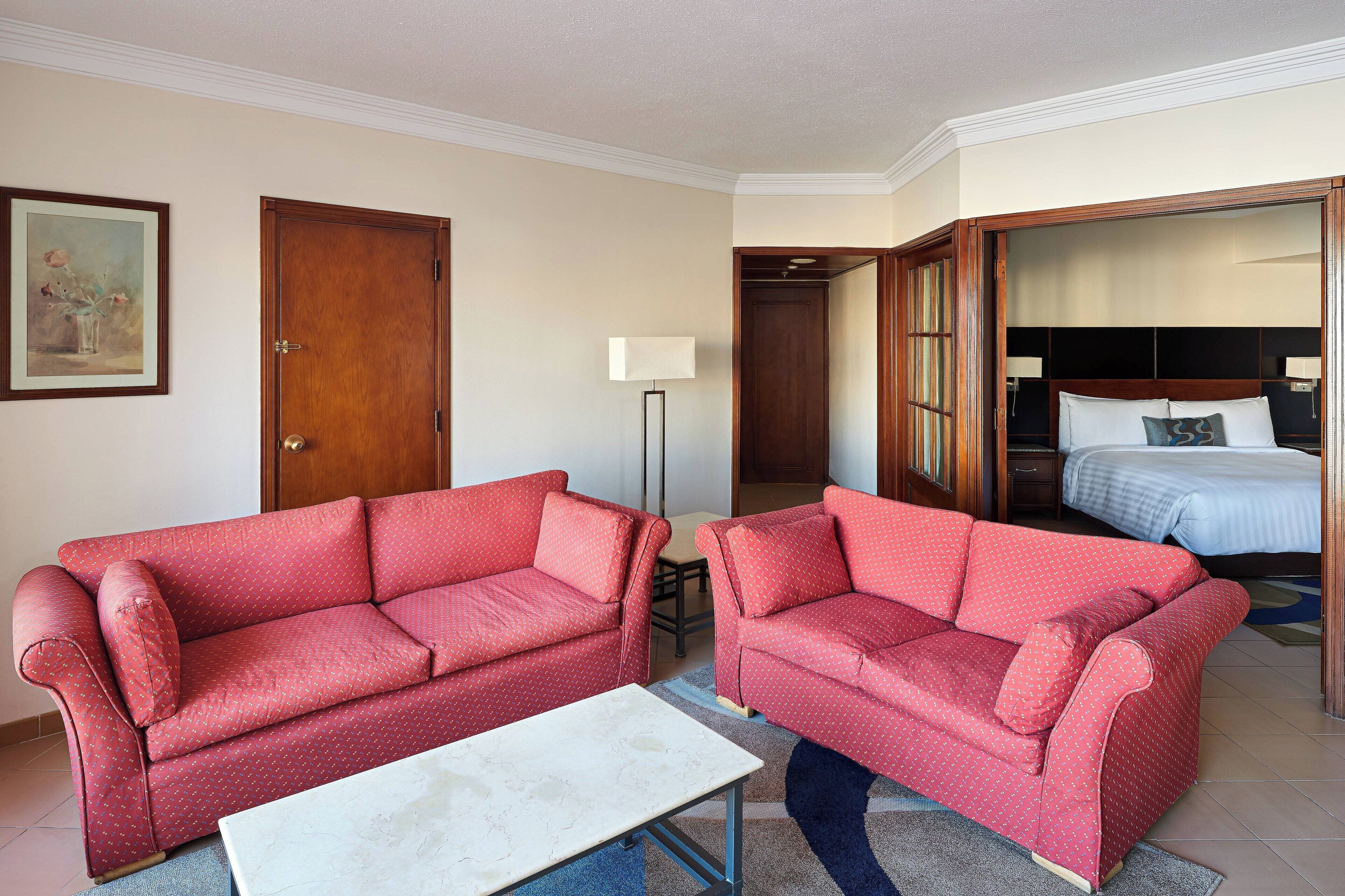 A hotel room with two red sofas, a white coffee table, a bed with a blue headboard, and a painting on the wall.