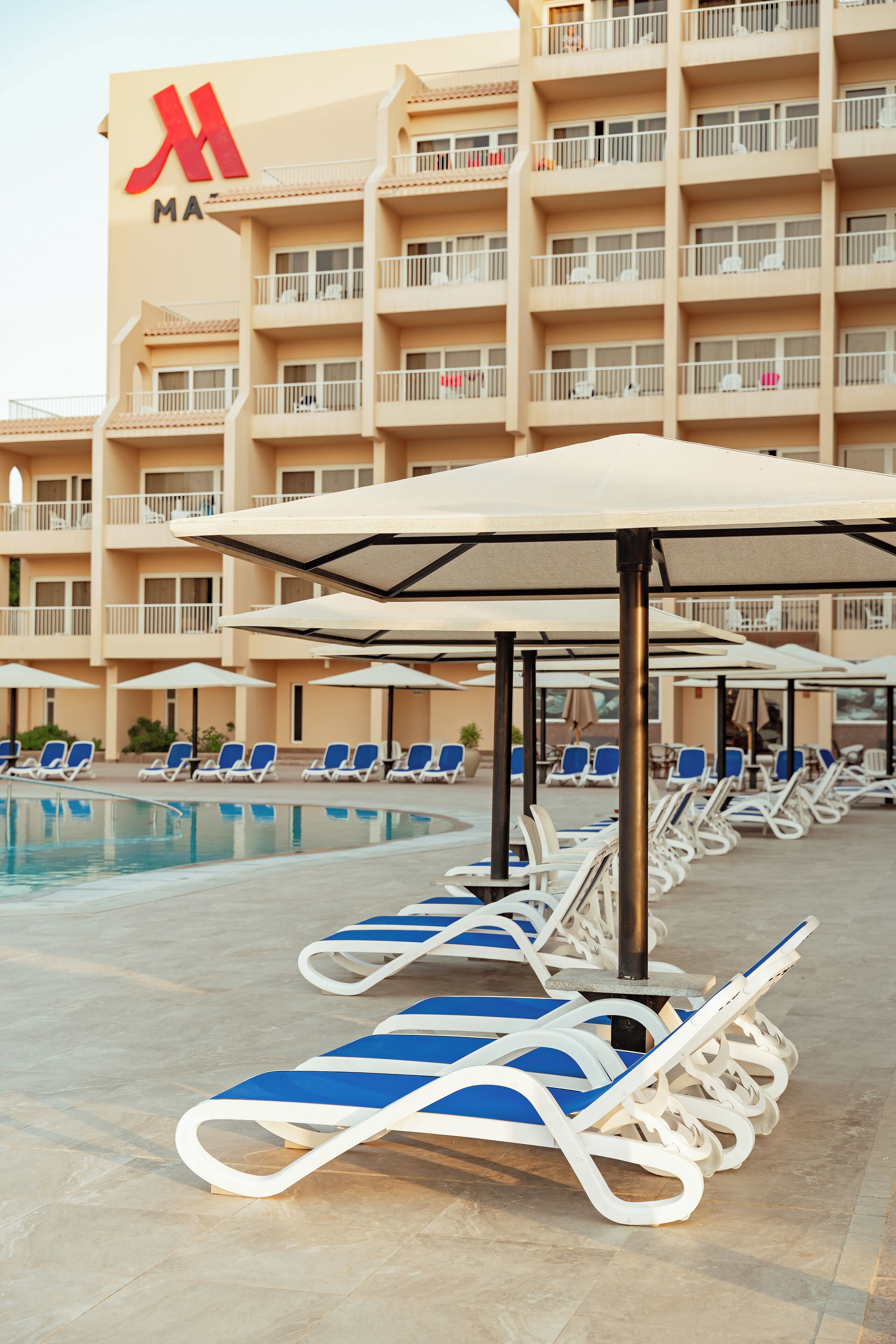 A hotel pool area with lounge chairs and umbrellas, a large building with balconies, and a clear sky.