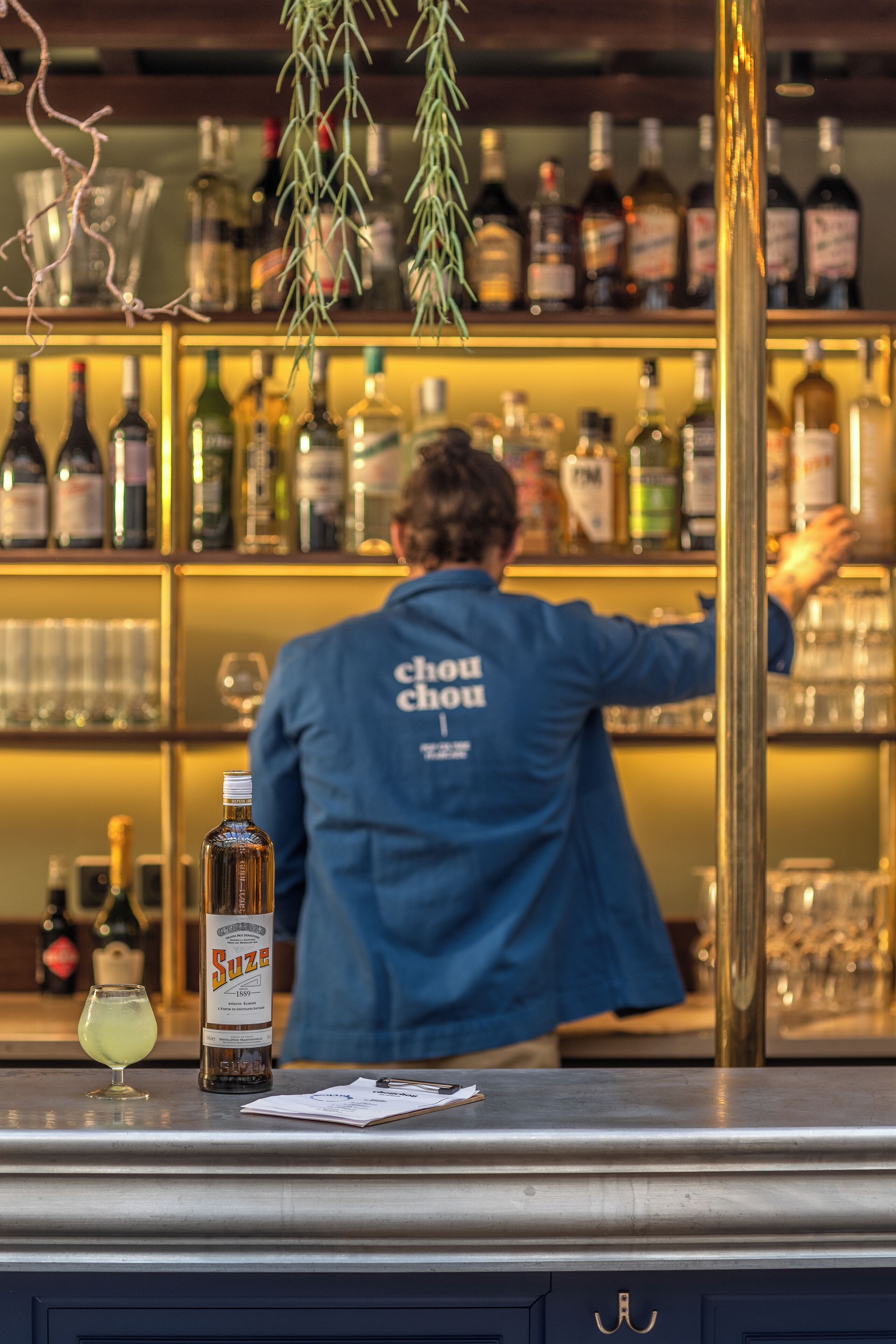 A bar area with a bartender in a blue jacket, shelves with various bottles, and a bottle of Suze wine on the counter.
