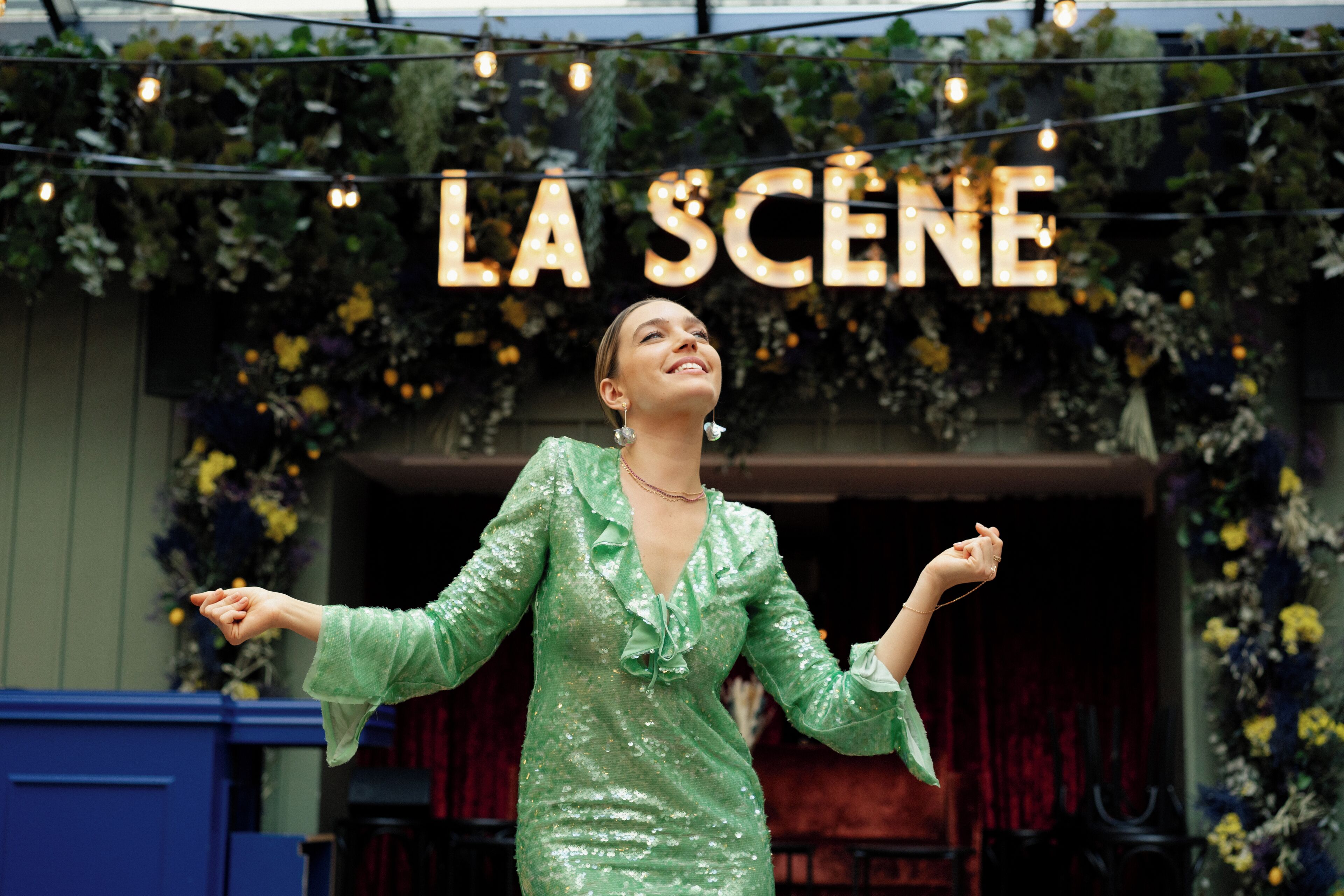 En person i en grön klänning som står framför en skylt med texten ”LA SCENE”.