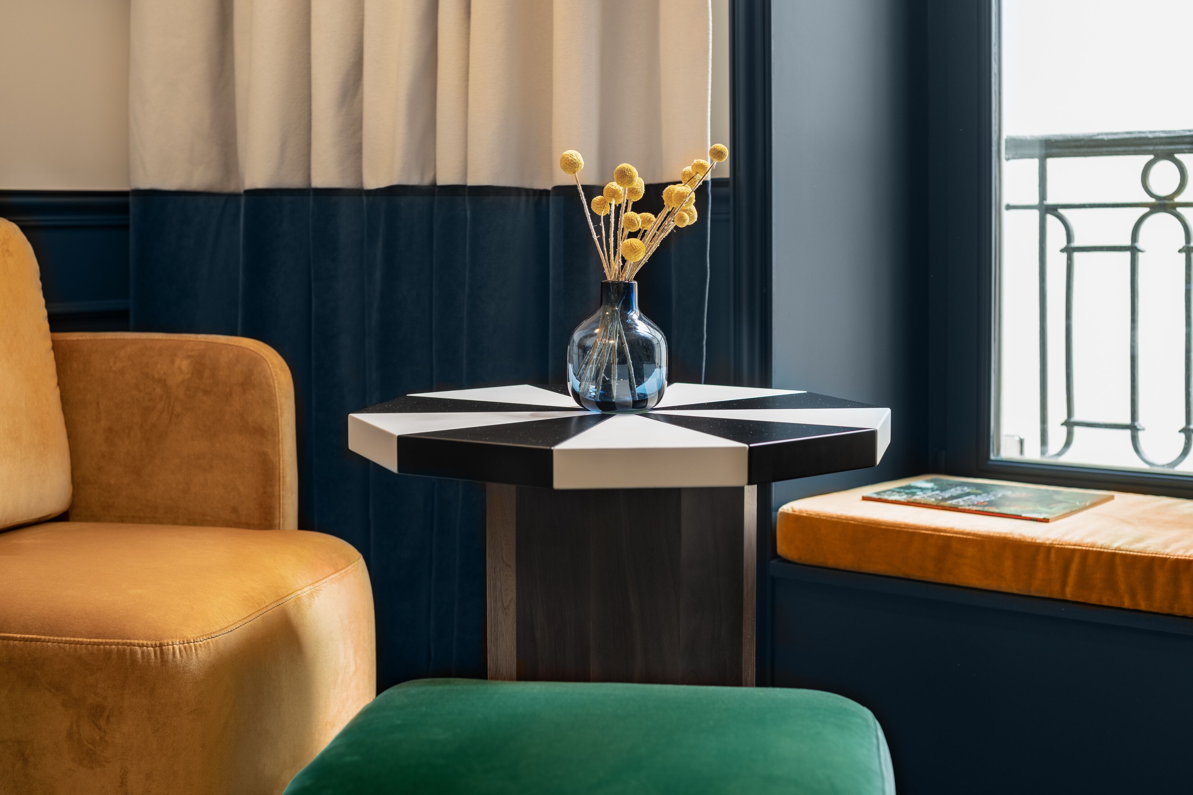 Une pièce avec un fauteuil en cuir marron, une petite table ronde ornée d’un vase de fleurs jaunes et une fenêtre donnant sur un balcon.
