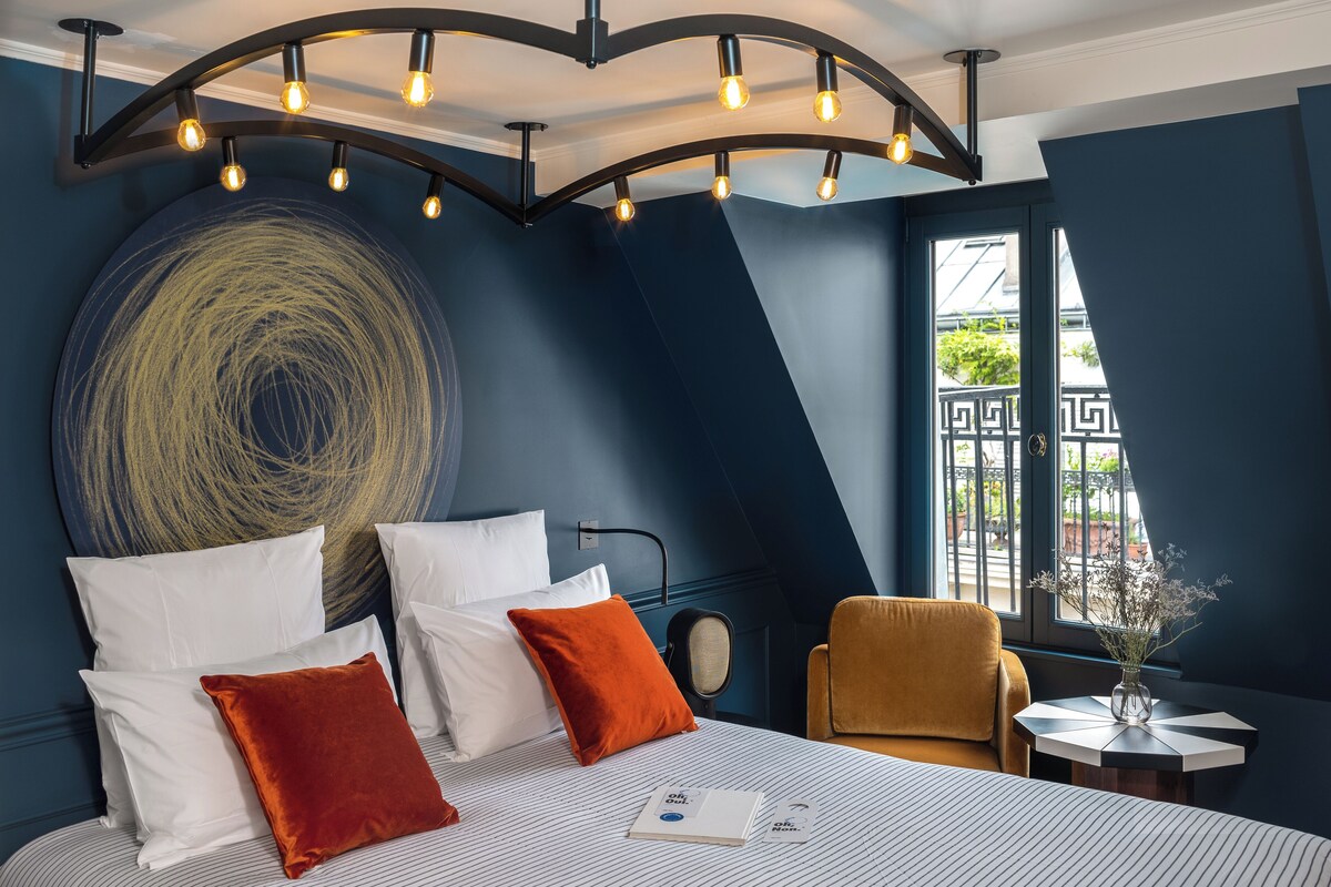 A modern hotel room with a large bed, a round wall art piece, a window with a view, and a unique ceiling light fixture.
