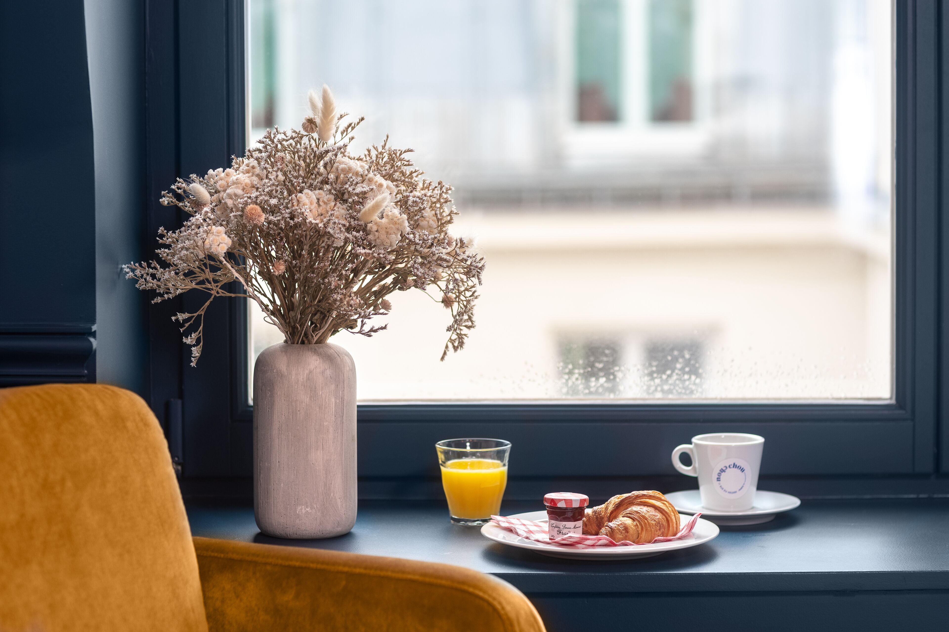 Una mesa para el desayuno con un jarrón de flores secas, un vaso de zumo de naranja, un croissant y una taza de café en un alféizar.