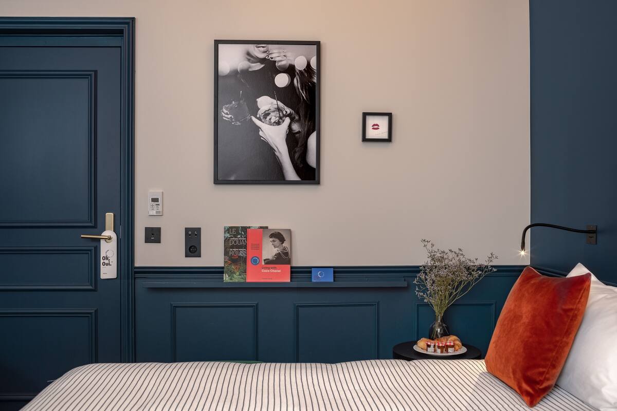 Una habitación de hotel con una cama, un cuadro enmarco, una mesita con un jarrón y velas, y una puerta con manija.