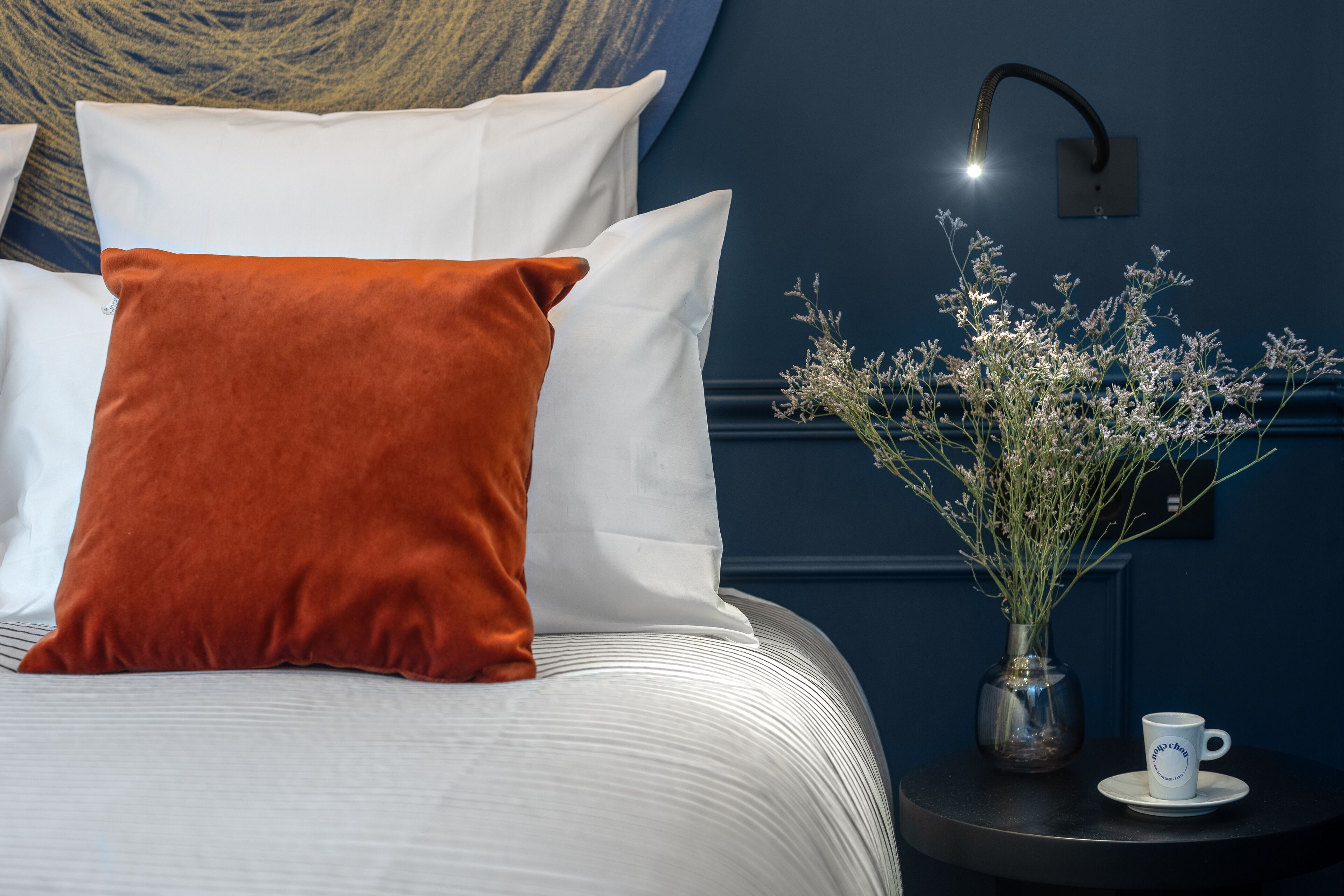 A neatly made bed with white and orange pillows, a vase with flowers, and a cup on a small table beside the bed.