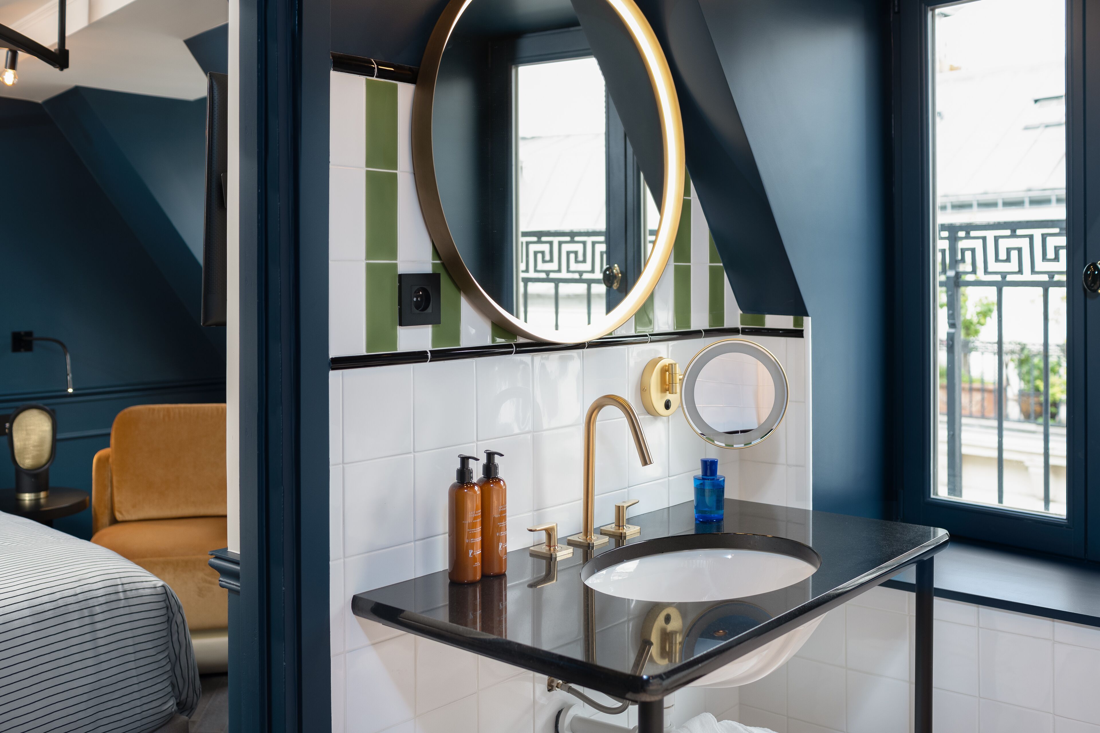 Une salle de bain moderne avec un lavabo noir, des robinets dorés et deux miroirs. On y trouve une fenêtre donnant sur un balcon et une porte ornée d’un motif géométrique.