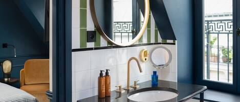Une salle de bain moderne avec un lavabo noir, des robinets dorés et deux miroirs. On y trouve une fenêtre donnant sur un balcon et une porte ornée d’un motif géométrique.
