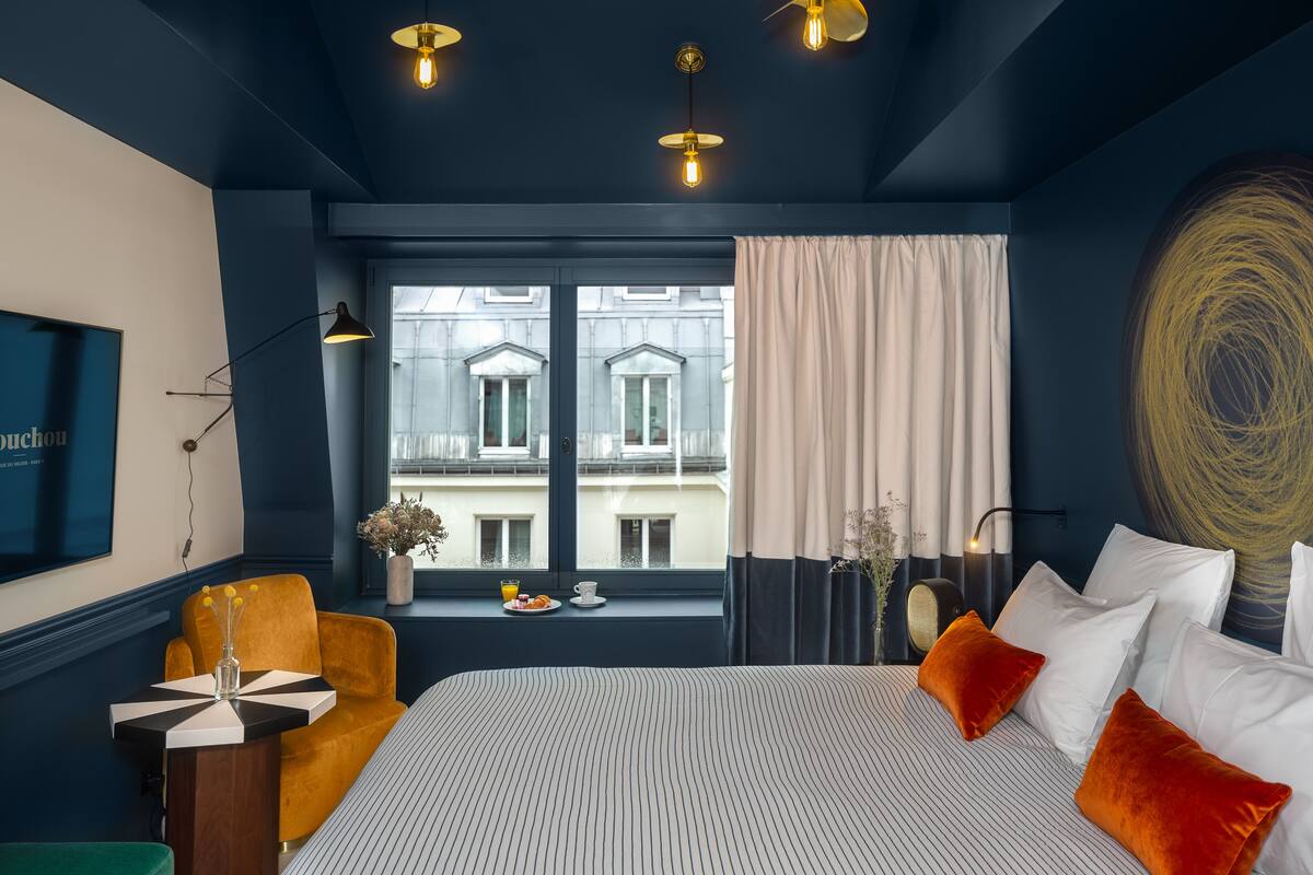 Habitación de hotel con una cama grande, vista de edificios a través de una ventana, un escritorio con una silla y un florero con flores.