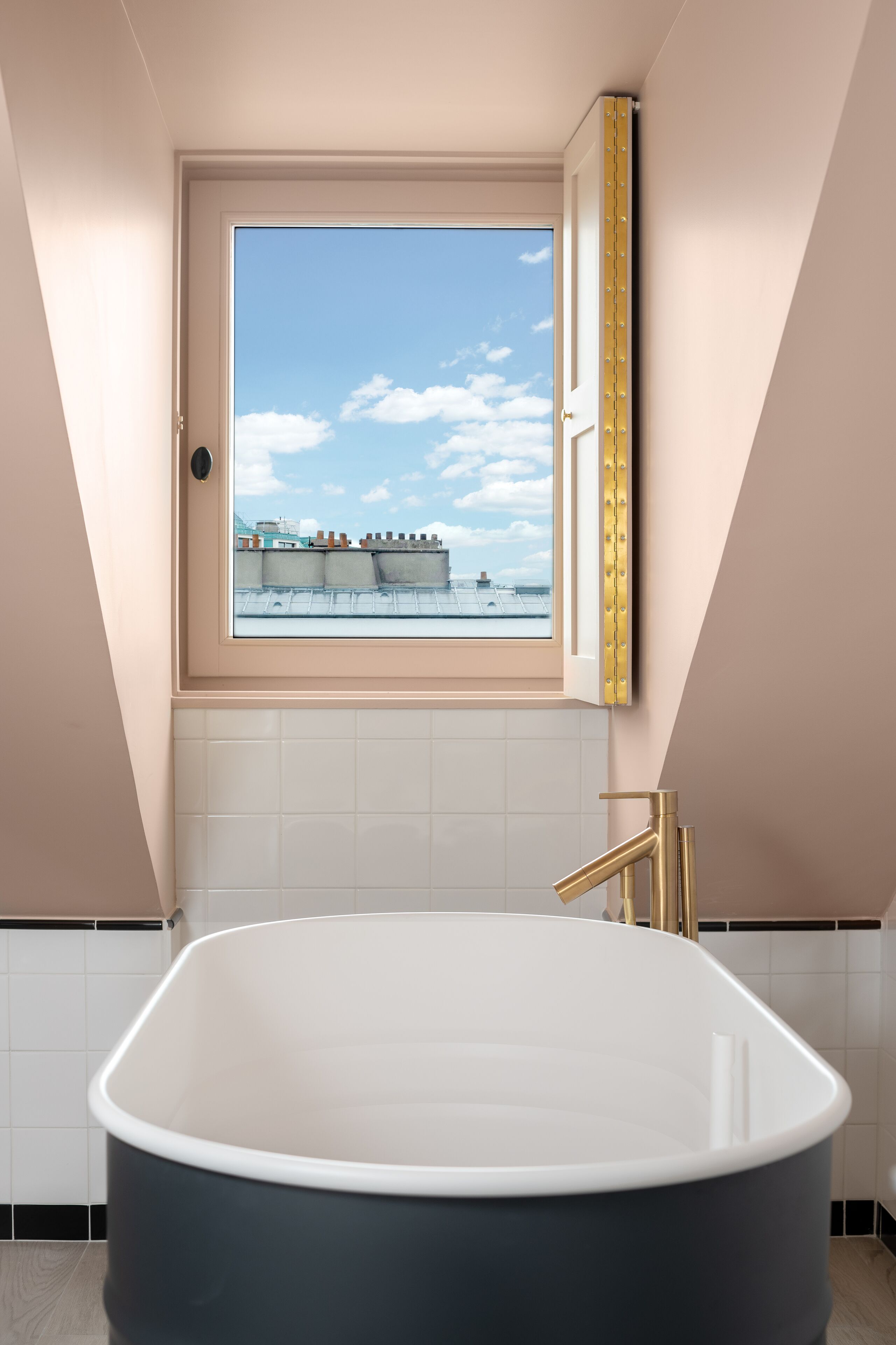 A modern bathroom with a freestanding bathtub, a window with a view of buildings, and a gold-framed mirror.