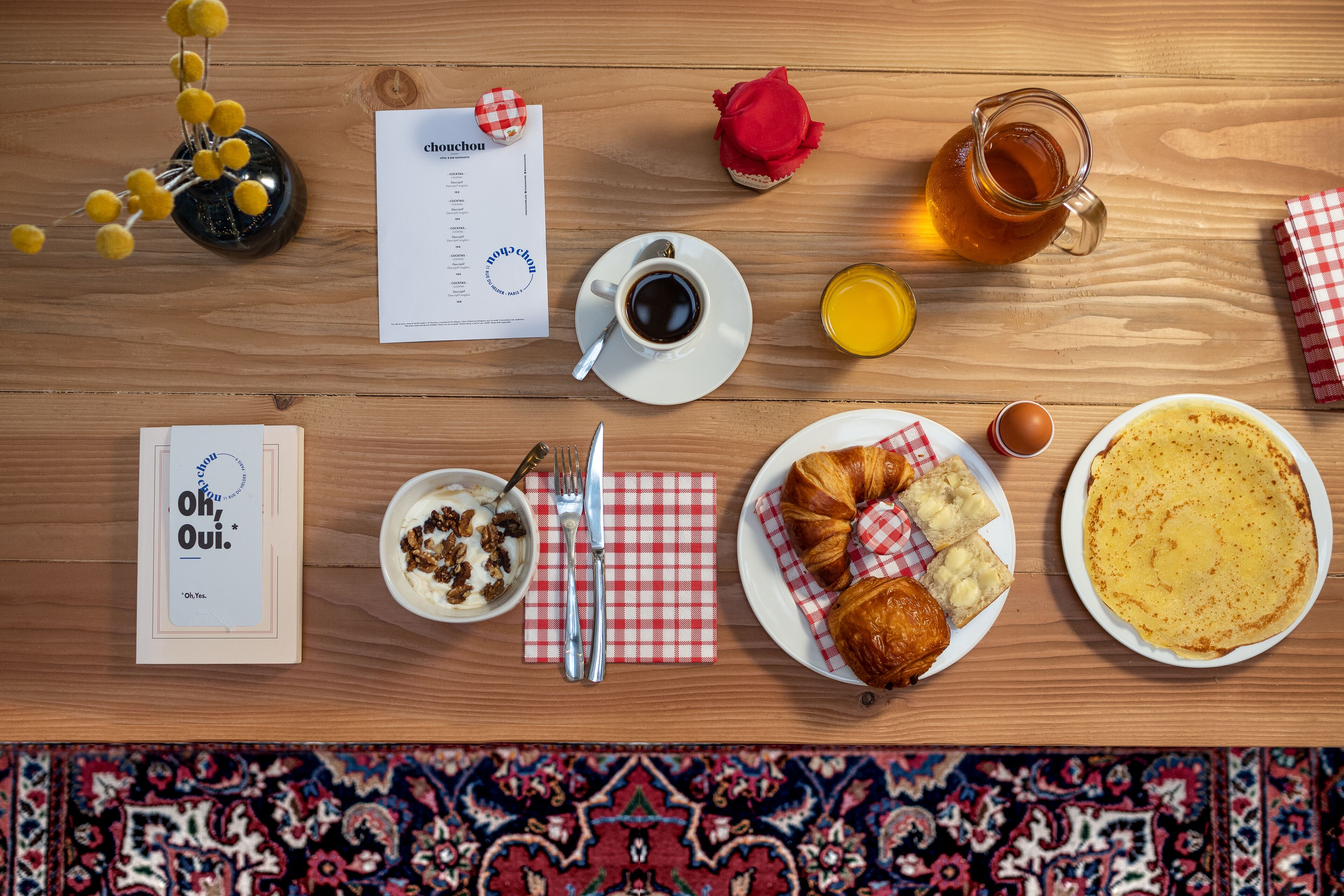A breakfast table with a croissant, a plate of pancakes, a cup of coffee, a glass of orange juice, a jar of honey, a red rose, a menu, and a book titled "Oh, Oui."