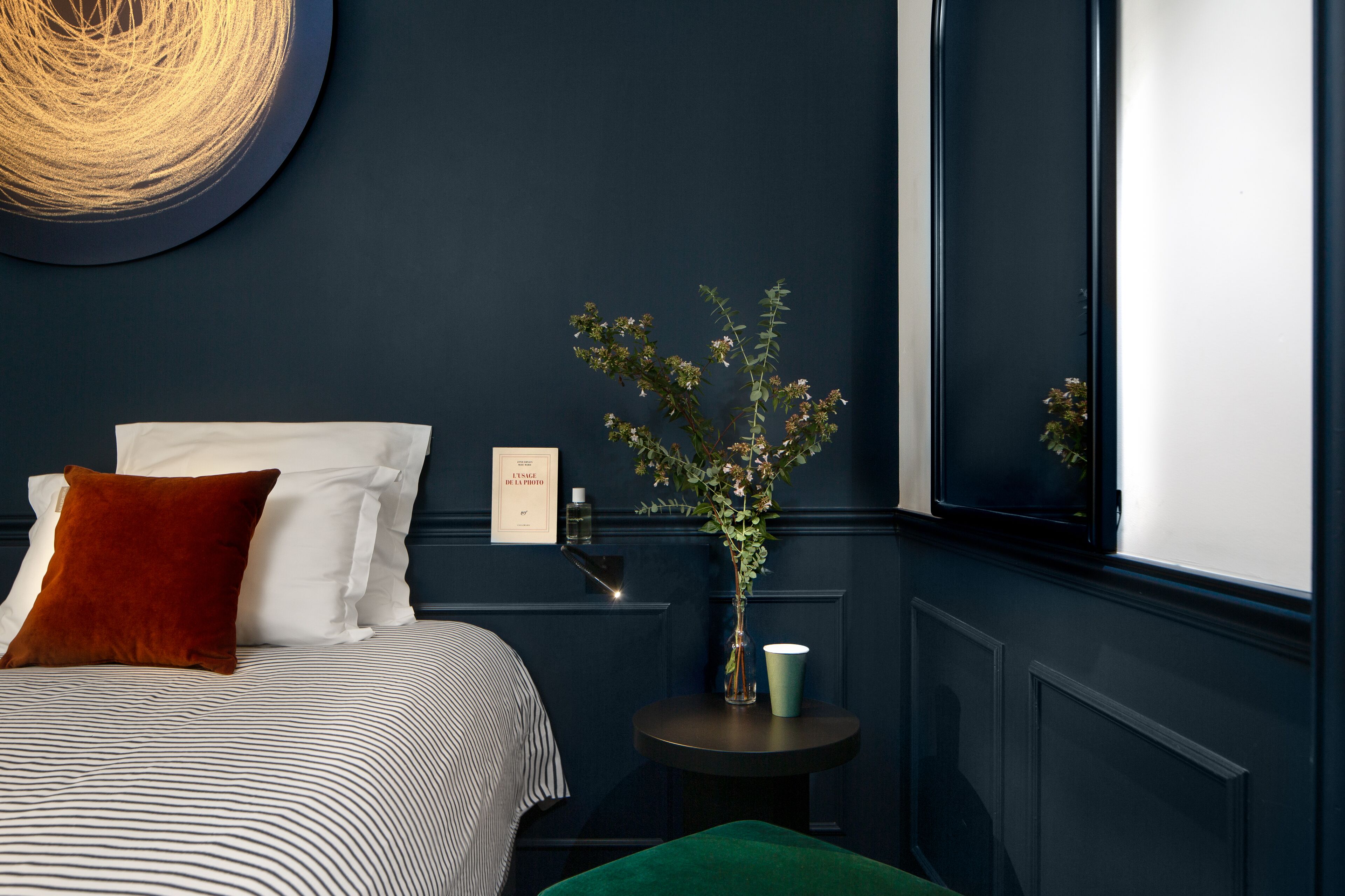 Un dormitorio con cama, almohadas, una mesita de noche con un jarrón y una taza, un reloj de pared y una ventana con vistas.