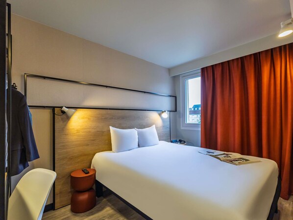 A hotel room with a large bed, a bedside table, a chair, a window with curtains, and a headboard with a light fixture. - ibis Metz Centre Cathédrale (Metz)