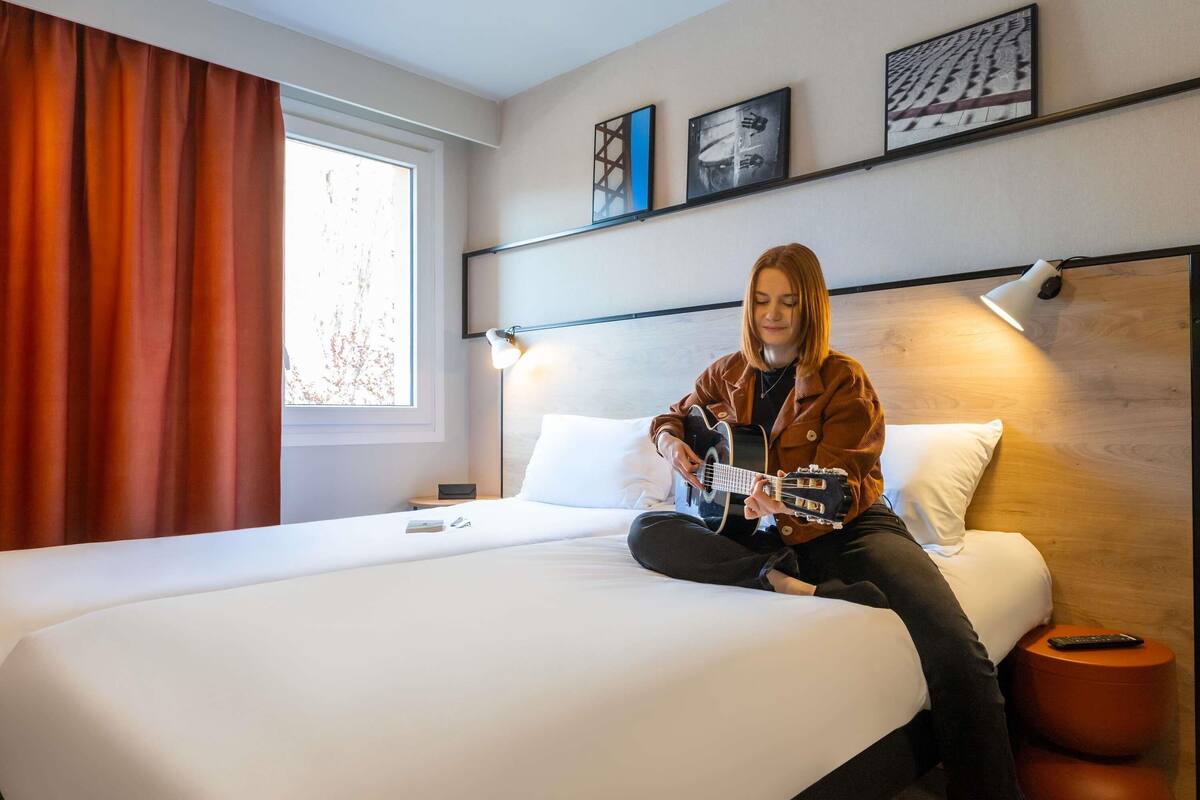 Quarto de hotel com uma cama, um violão, uma luminária e obras de arte na parede.