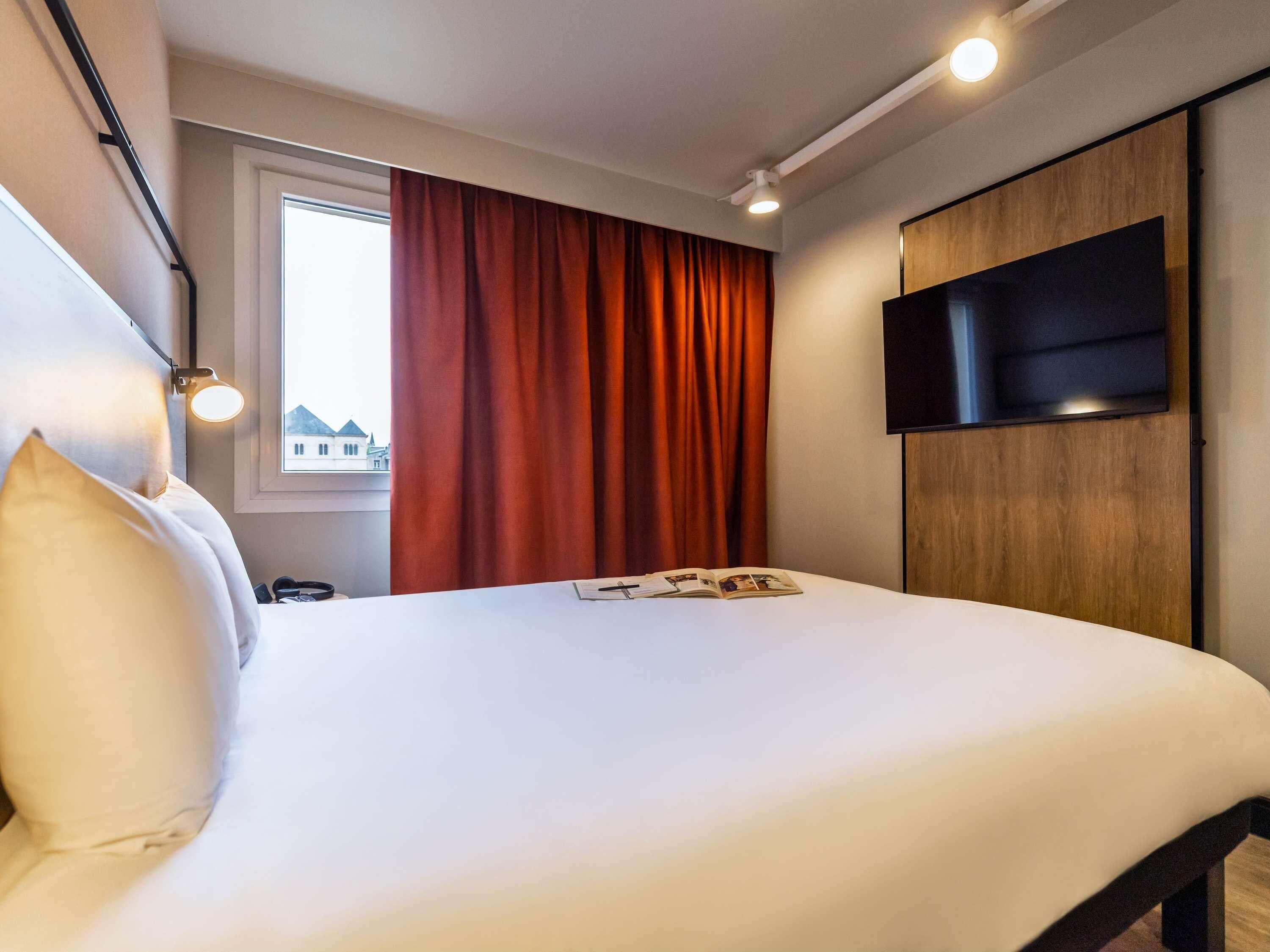 Hotellihuone, jossa on sänky, ikkuna verhoilla, seinälle asennettu televisio ja yöpöytälamppu.